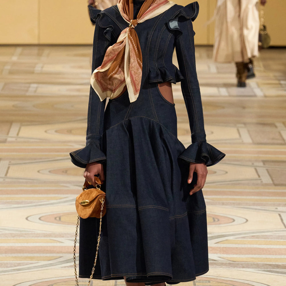 Model walking on a runway wearing a dark blue outfit with a beige scarf.