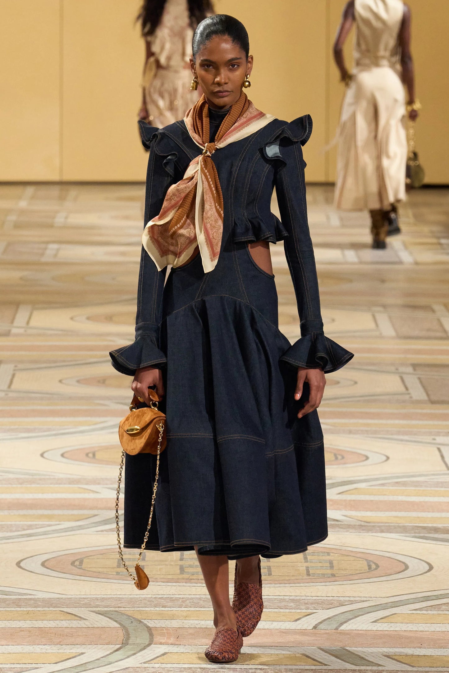 Model walking on a runway wearing a dark blue outfit with a beige scarf.