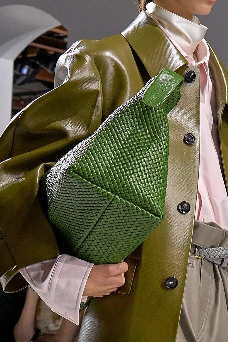 Model walking on a runway wearing a green coat and beige skirt, holding a green bag.
