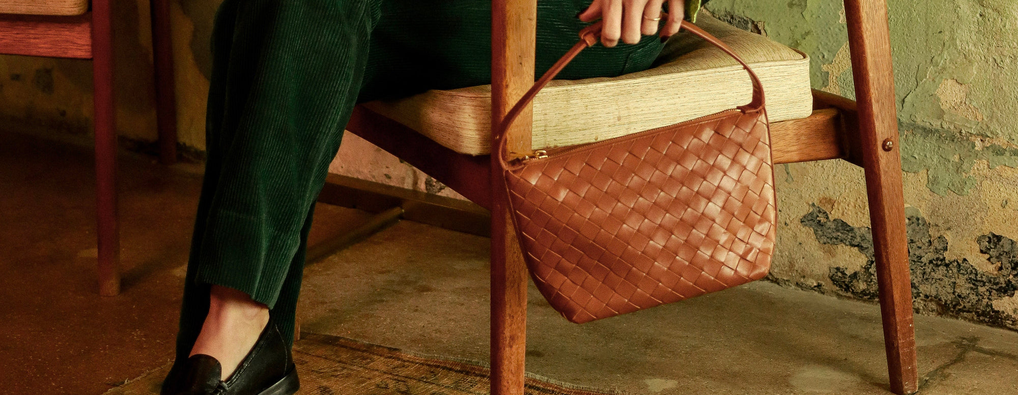 Person sitting on a wooden chair holding a brown leather bag in a room with peeling paint.