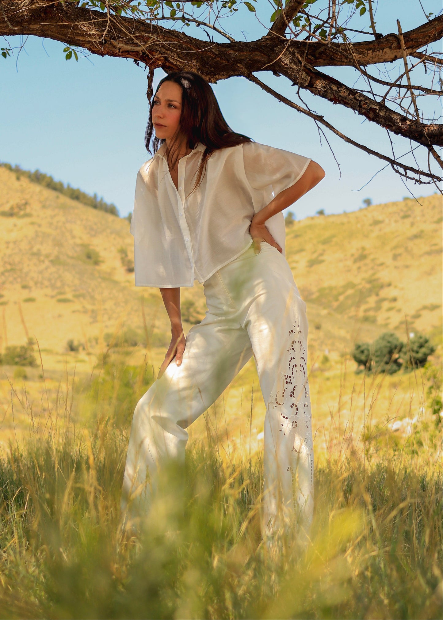 A woman stands outdoors in a grassy field under a tree, wearing a loose white short-sleeve top and matching white wide-leg pants with black script text down one leg.