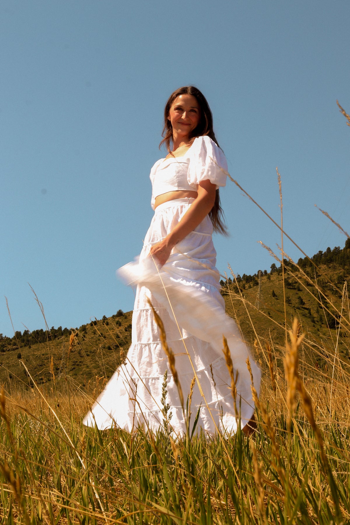A woman wearing a flowing white two-piece outfit stands in a grassy field under a clear blue sky, with hills in the background.