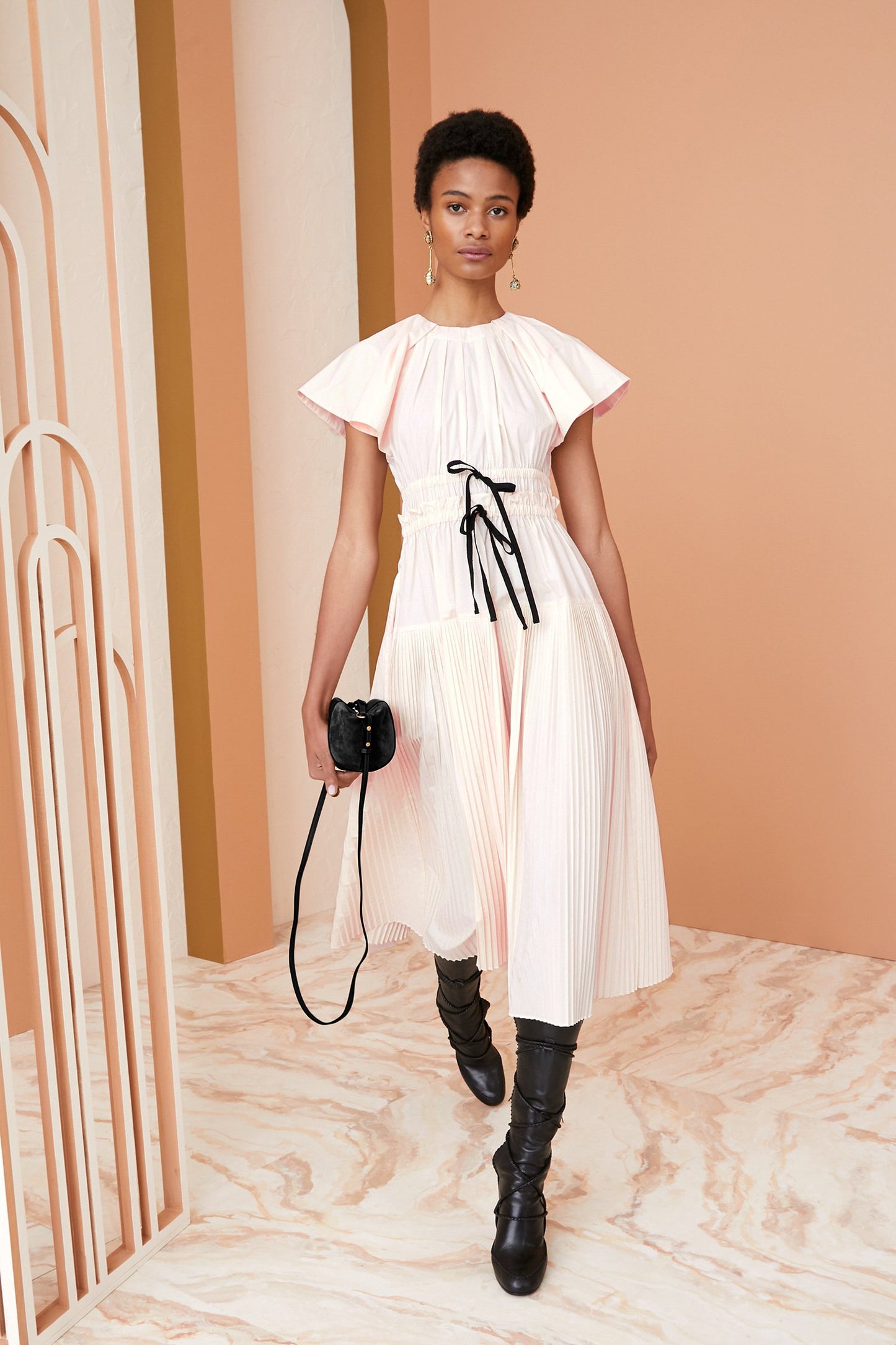 Woman in a white dress with black boots holding a camera against a peach-colored wall.