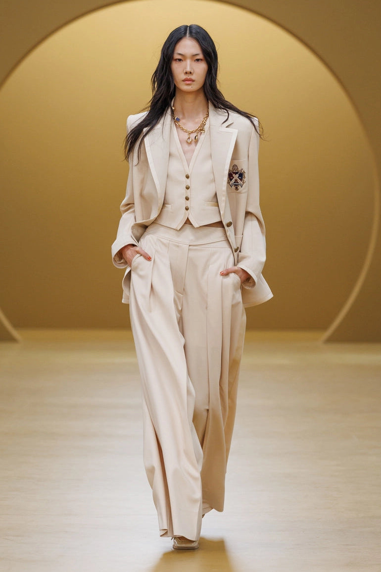 A model walks down a runway wearing a light beige tailored suit with a vest, matching wide-leg trousers, and a structured blazer. The background features a minimalist warm beige studio setting with a large circular architectural element.