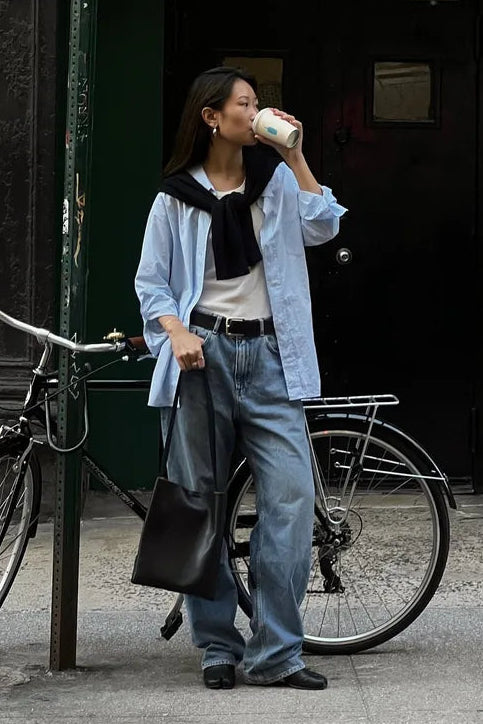 A woman stands on a city sidewalk in front of a dark doorway, holding a paper coffee cup. She is wearing a light blue button-up shirt over a white top with a black neck scarf, light-wash wide-leg jeans, and black shoes.