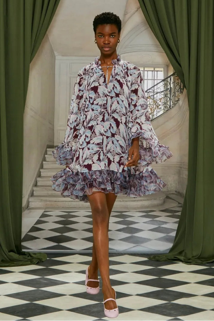 Woman in a floral dress standing in a grand hall with green curtains and checkered floor.