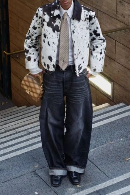 A model stands on outdoor concrete stairs wearing a black and white cow-print jacket, wide-leg dark jeans, a beige patterned tie, a white shirt, and a black bow headband.