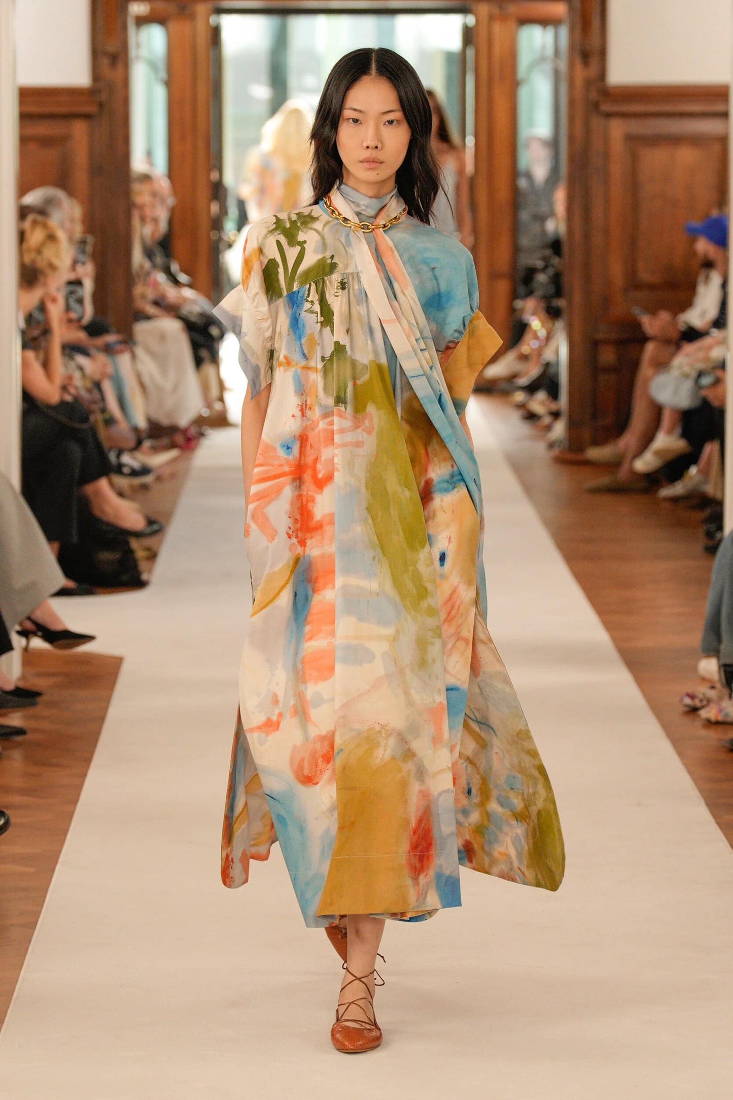 Model walking down a runway wearing a colorful dress with a patterned scarf.